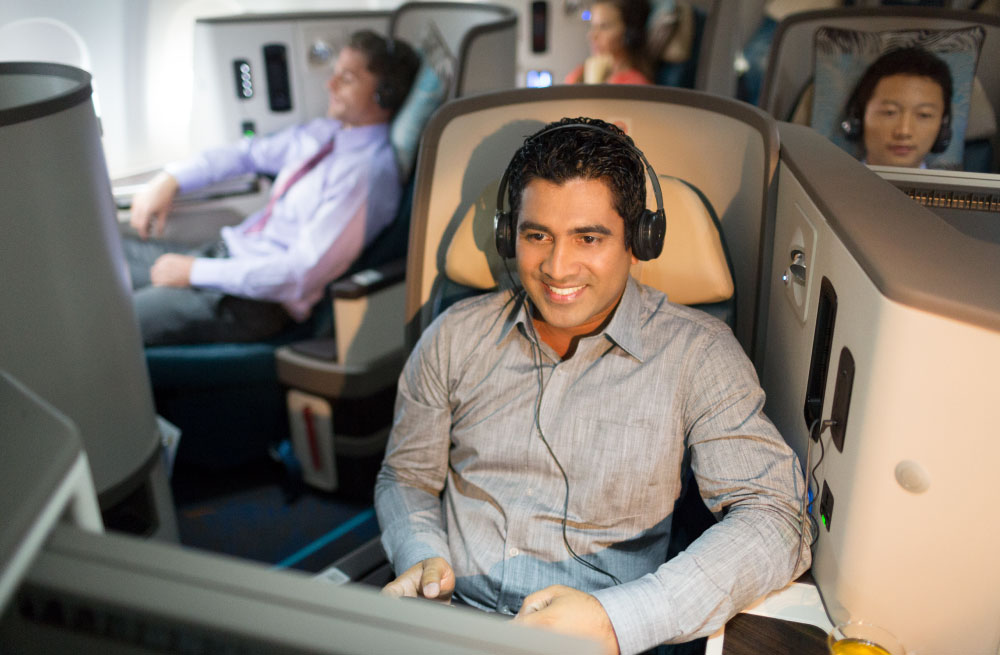 Female in business class wearing headphones looking at the screen in front of her with an open-mouthed smile