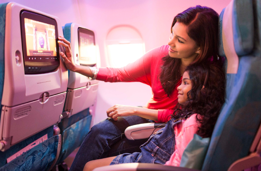 A male passenger with headphones on reclines in seat pressing a button on the entertainment system in front of him